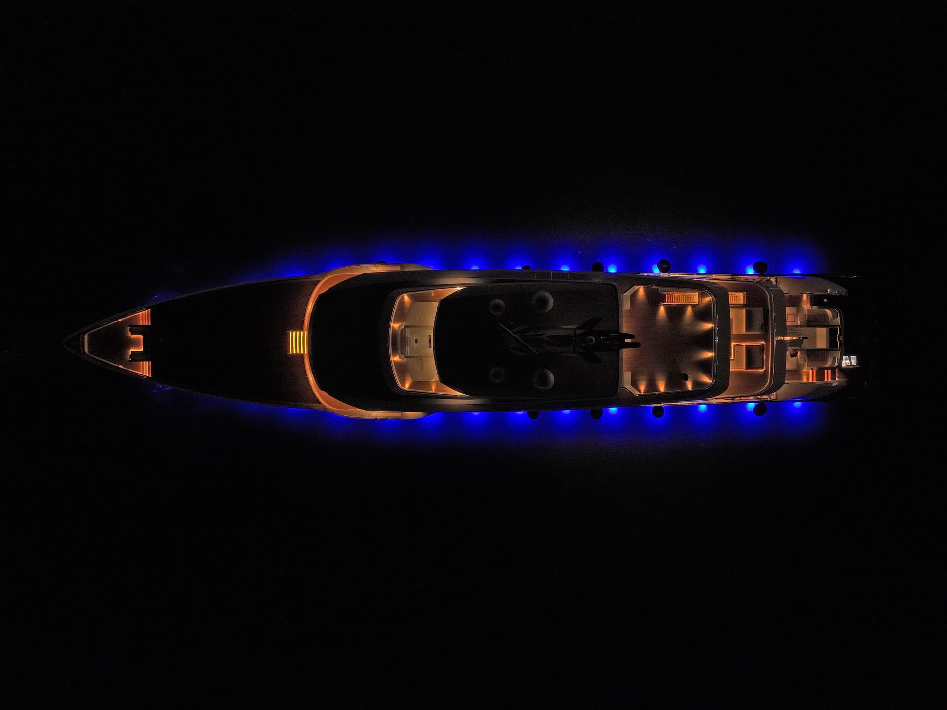 Aerial top-down view of the 78-meter Admiral yacht at night, illuminated by warm interior deck lighting and a striking blue glow from its underwater lights against a black background.