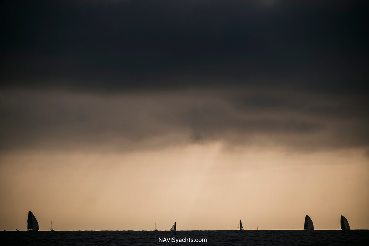 A tight shot of a solo sailor, like Justine Mettraux, at the helm of their yacht, their face showing a mix of intense focus and exhaustion while navigating the challenging "lonely latitudes."
